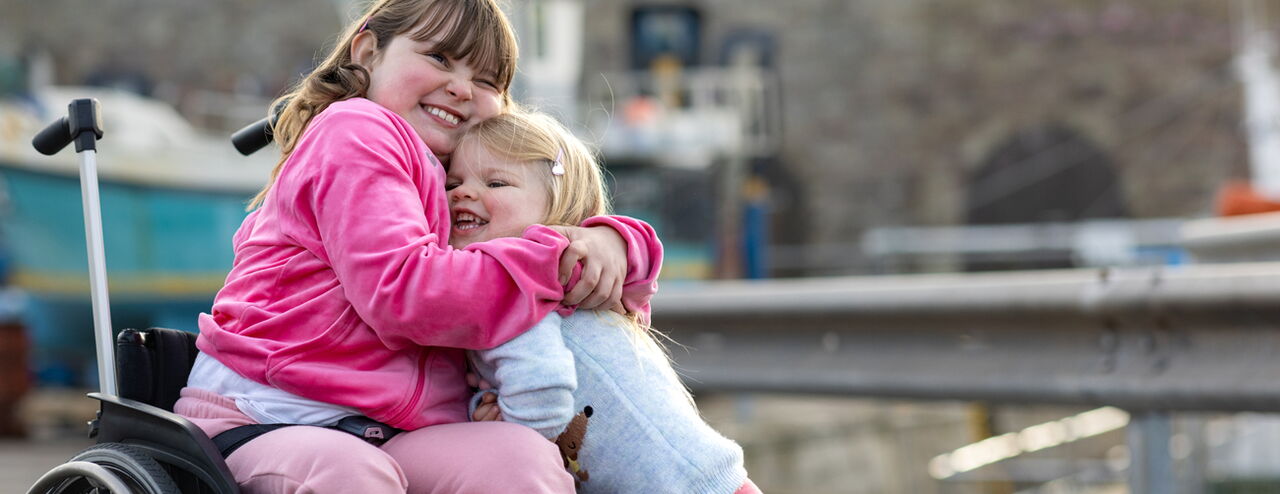 Eine Person im Rollstuhl trägt einen pinken Hoodie und umarmt ein Kind, das einen hellblauen Pullover trägt. Im Hintergrund sind Boote und eine Mauer sichtbar.