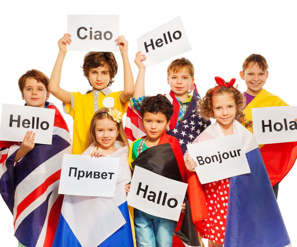 Neun Kinder halten Plakate mit Begrüßungen in verschiedenen Sprachen: 'Ciao', 'Hello', 'Hola', 'Bonjour', 'Hallo', 'Привет'. Sie tragen bunte Kleidung und Umhänge mit Flaggen.