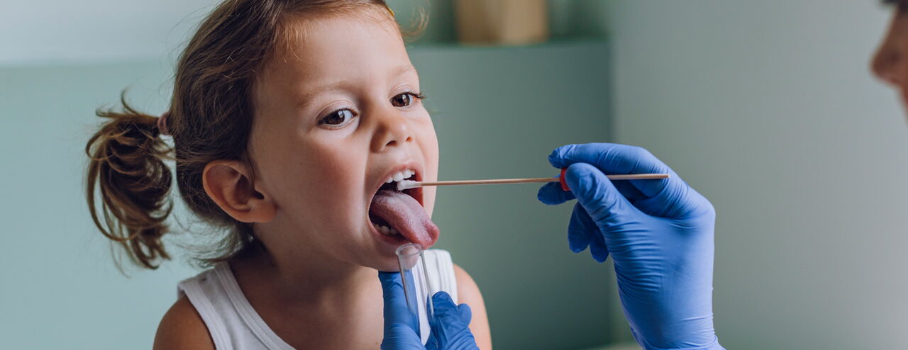 Ein Kind mit einem Zopf trägt ein weißes Oberteil. Eine Hand mit blauen Handschuhen hält einen Wattestäbchen in Richtung des Mundes des Kindes.