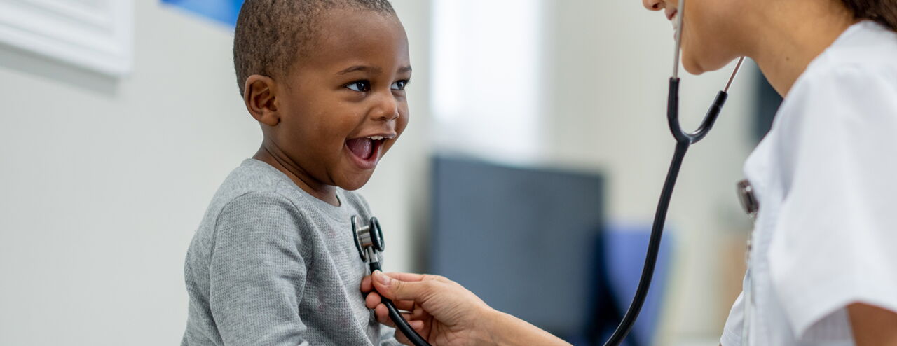 Ein Kind sitzt auf einem Stuhl und wird von einer medizinischen Fachkraft mit einem Stethoskop untersucht. Die Fachkraft trägt einen weißen Kittel.
