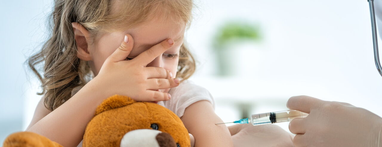 Ein Kind hält einen braunen Teddybären und hat einen Arm zur Seite gedreht, während eine Hand mit einer Spritze eine Impfung verabreicht.